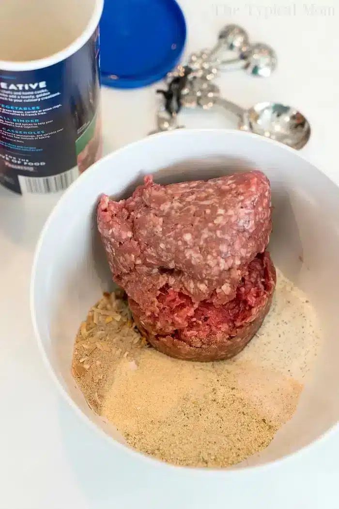 A bowl containing ground meat and various spices ready to become a good Ninja Foodi meatloaf. There's a container in the background and a set of measuring spoons.
