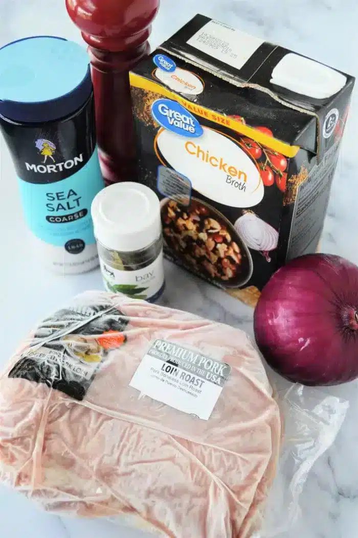 A selection of ingredients for Ninja Foodi pork recipes is displayed, featuring a package of pork loin roast, a container of coarse sea salt, a jar labeled "bay," a carton of chicken broth, a red onion, and a pepper grinder.
