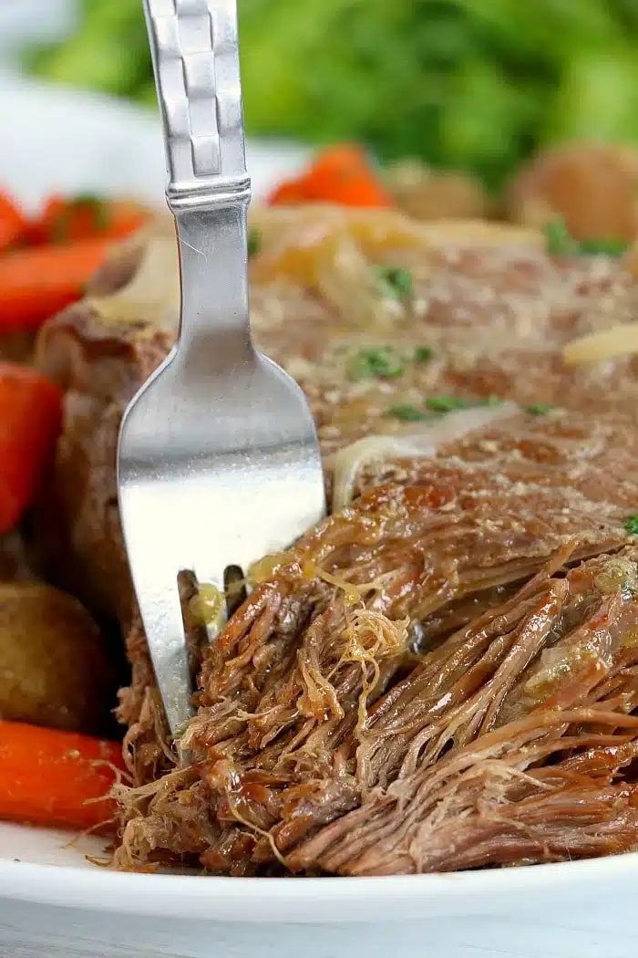 A fork shredding tender pot roast with carrots and potatoes on a plate, greens in the background—one of our favorite Ninja pressure cooker recipes.