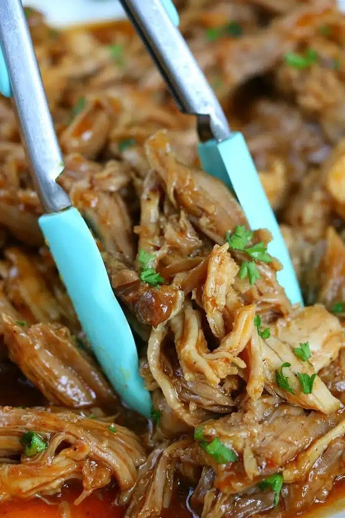 Tongs holding shredded, saucy pulled pork garnished with chopped parsley—solid for trying out new ninja pressure cooker recipes.