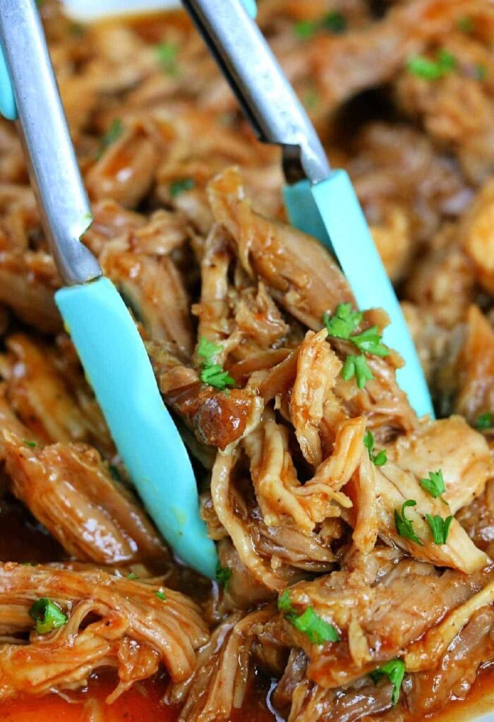 Tongs holding shredded barbecue chicken garnished with chopped parsley, served alongside tender Ninja Foodi pulled pork.