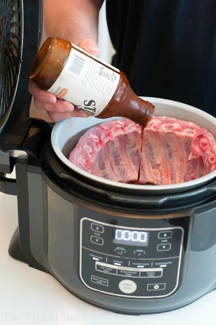 Person pouring barbecue sauce onto raw ribs in a Ninja Foodi, getting the Ninja Foodi ribs ready to be cooked to tender perfection.
