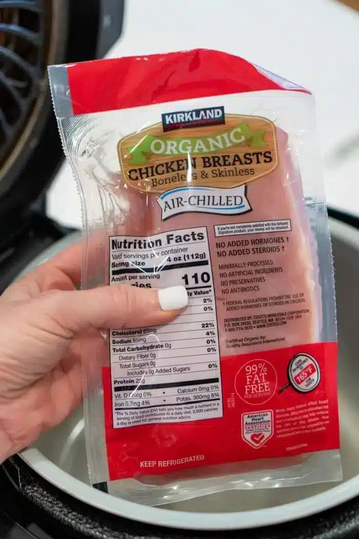 A package of Kirkland organic chicken breasts is held by a hand over a Ninja Foodi, ready to transform into good shredded chicken. The packaging, labeled as boneless, skinless, and air-chilled, displays nutritional facts and other details.