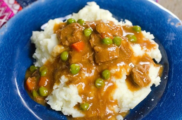Mashed potatoes topped with a savory ninja foodi stew, peas, and carrots rest elegantly on a blue plate.