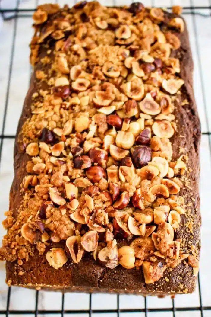 A loaf nutella bread topped with a crumbly mix of chopped nuts, including hazelnuts, reminiscent of Nutella bread, sits on a cooling rack. The golden-brown cake has a textured top with scattered pieces of nuts and crumbs visible.