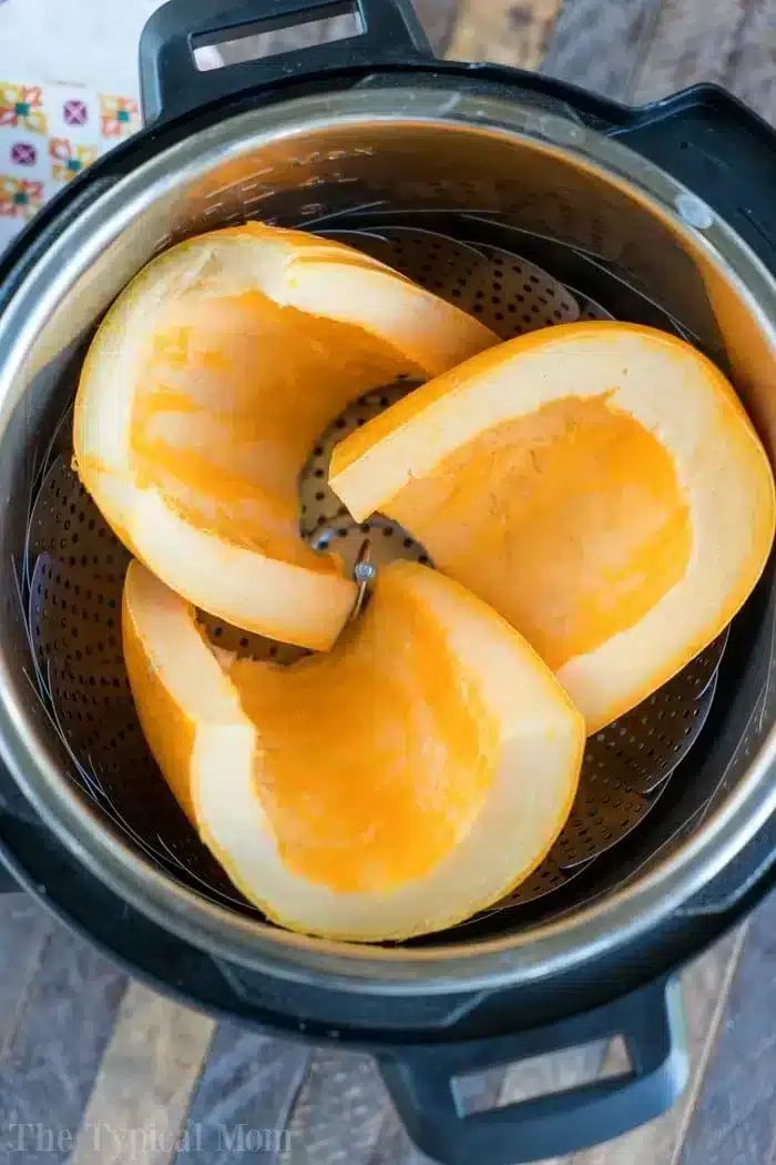 Three pumpkin slices rest inside an Instant Pot with a steamer basket, solid for October's cozy recipes. Cut into large wedges, they showcase their bright orange flesh. The pot sits invitingly on a wooden surface.