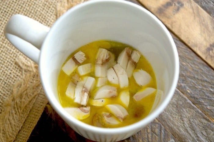 A white mug filled with a eggs and sliced mushrooms, reminiscent of an omelette in a mug, rests on a wooden surface.