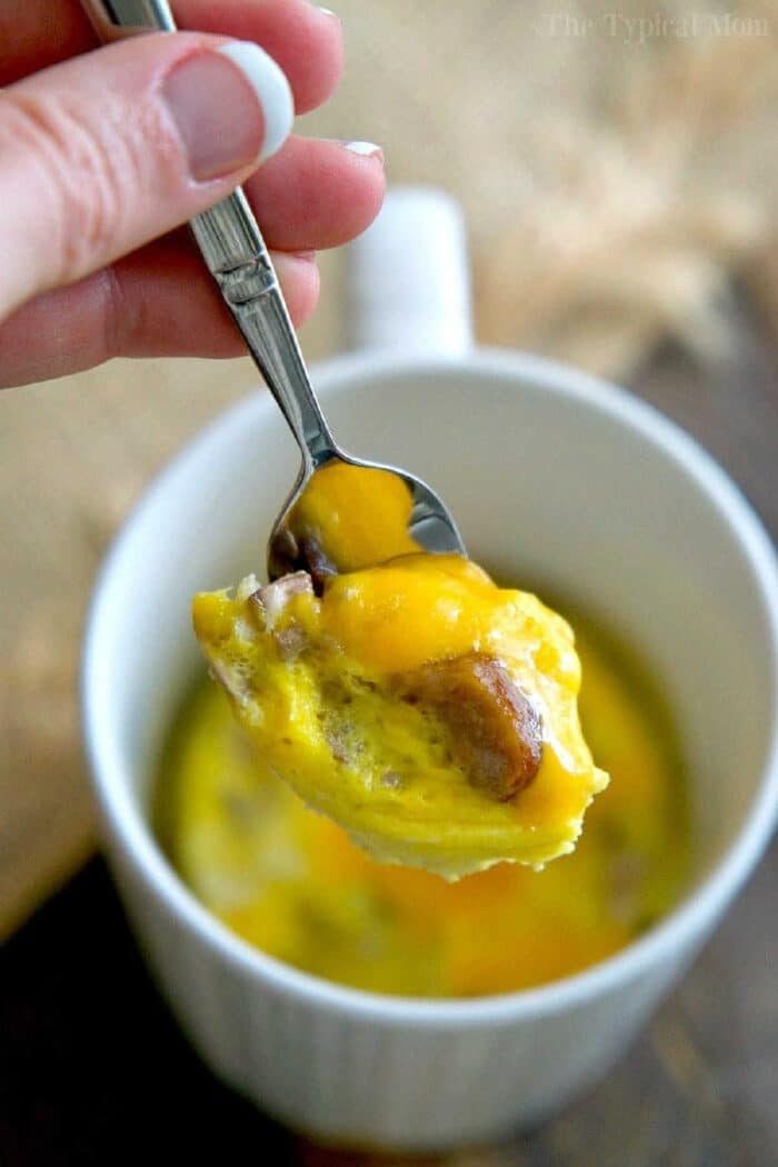 A hand holds a spoon with cheesy egg and sausage above a white mug filled with omelette in a mug.