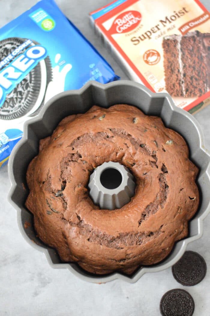 An Oreo Cake with cake mix sits elegantly in a pan, surrounded by Oreo cookies and cake mix boxes on a gray surface.