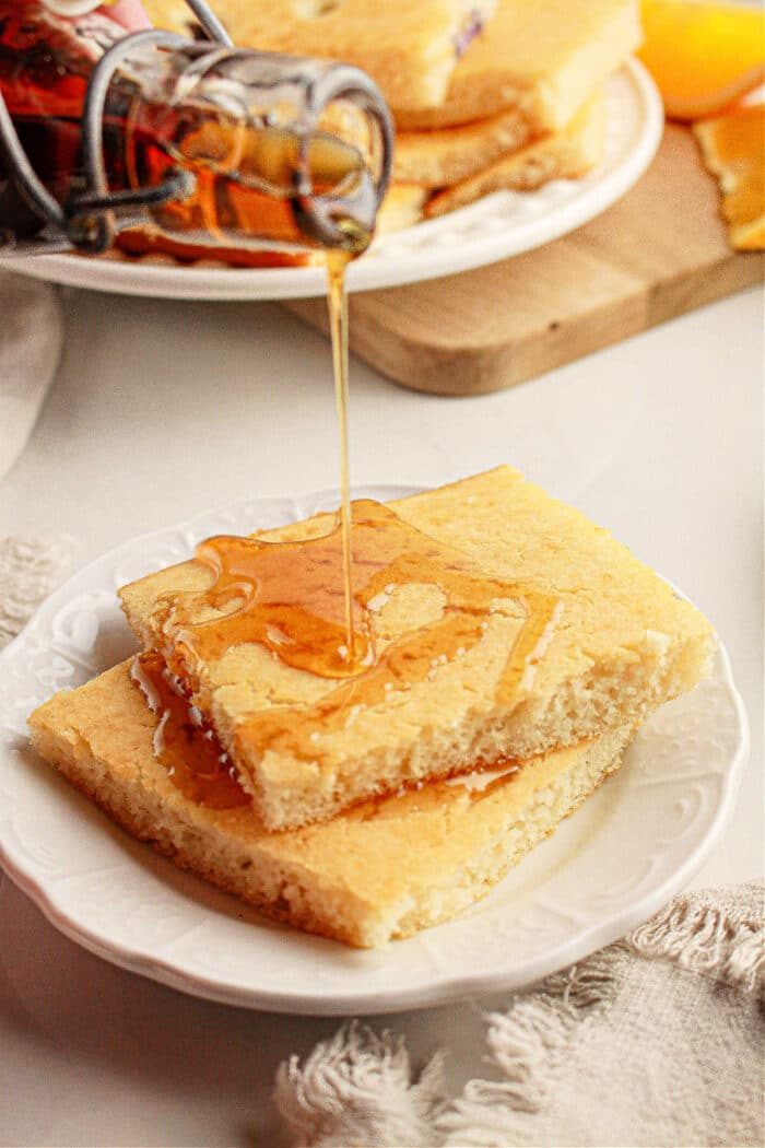 Pouring syrup over two stacked pieces of cornbread on a white plate, with more oven baked pancakes with pancake mix in the background.