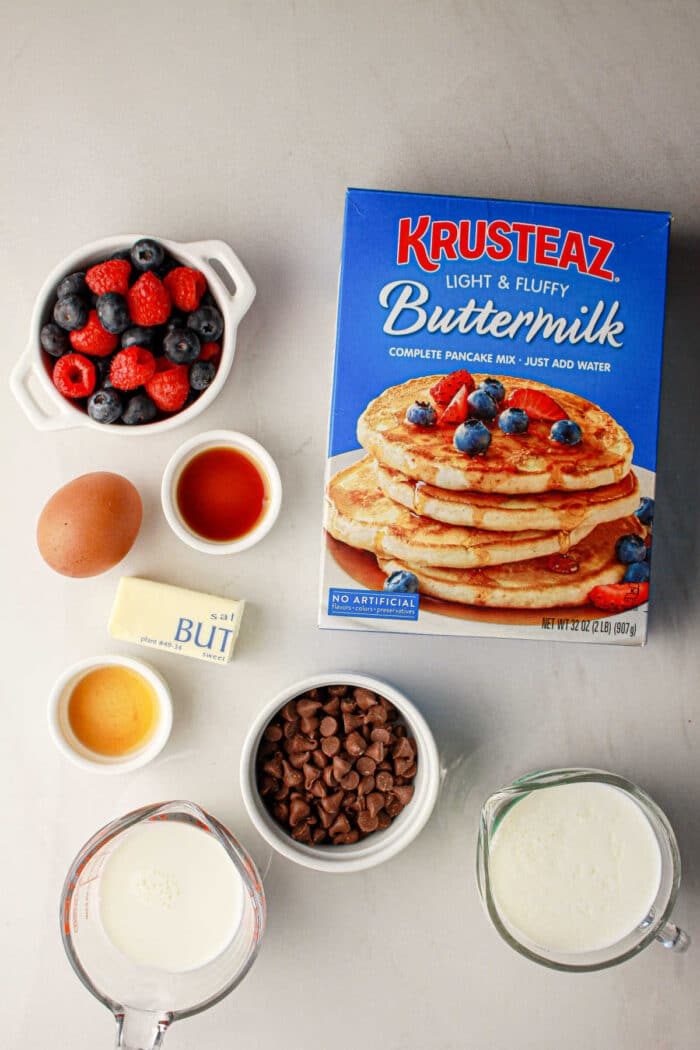 Box of pancake mix, berries, chocolate chips, butter, milk, egg, vanilla, and syrup on a gray surface—everything you need for good oven baked pancakes with pancake mix.