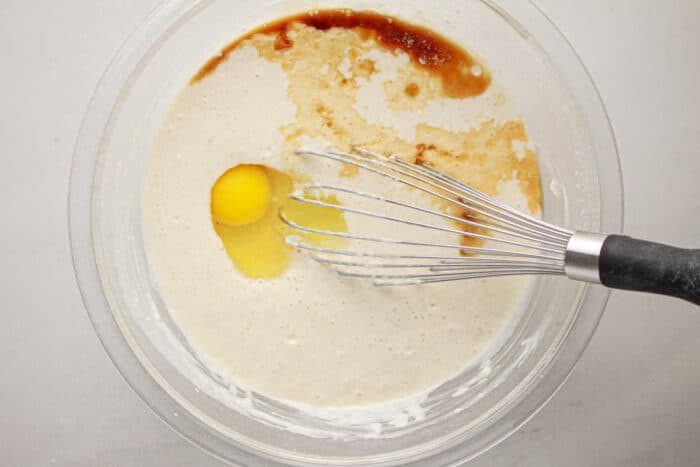 A whisk is in a bowl with batter, an egg, vanilla, and other ingredients being mixed together for oven baked pancakes with pancake mix.