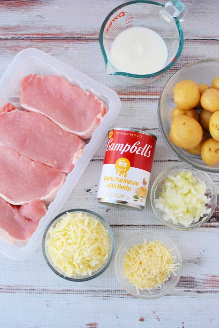 Raw pork chops with potatoes, chopped onions, shredded and grated cheese, milk, and a soup can are arranged on a table—solid ingredients for good oven baked pork chops.