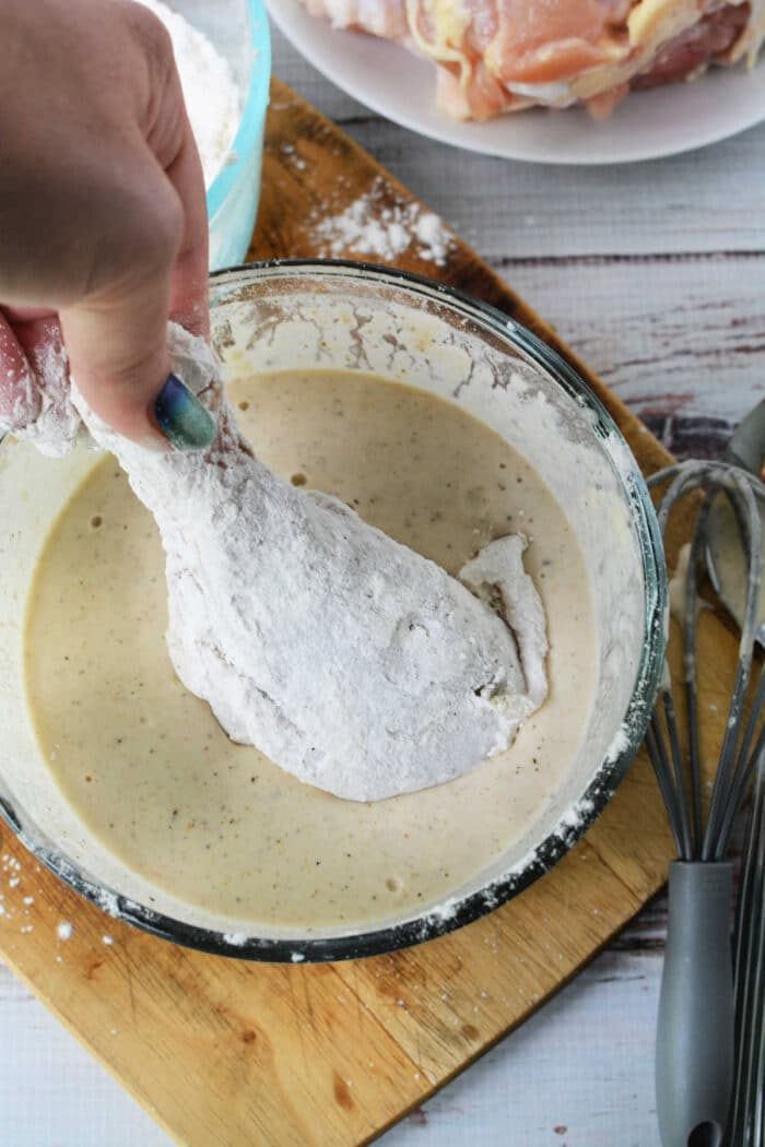 A hand dips a floured chicken drumstick into a bowl of batter on a wooden cutting board, preparing it for good pan fried chicken.