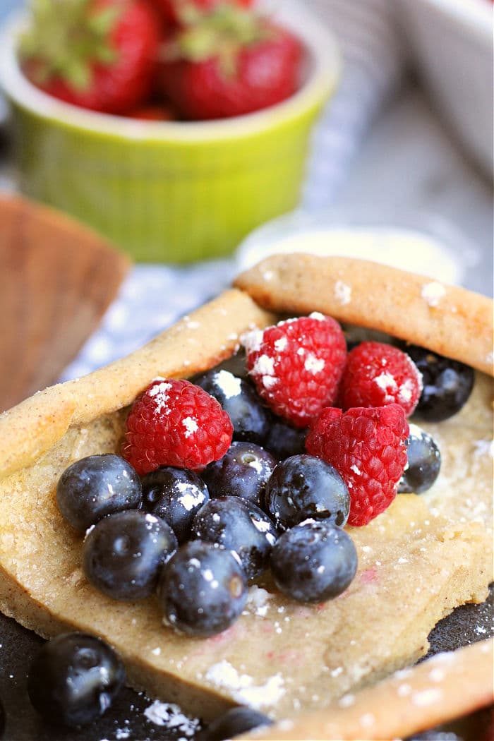 A close-up of a piece of pastry topped with powdered sugar, blueberries, and raspberries. In the background, there is a green bowl filled with fresh strawberries. The scene resembles a delightful setting where pancakes in the oven could be baking to perfection for a good dessert or snack.
