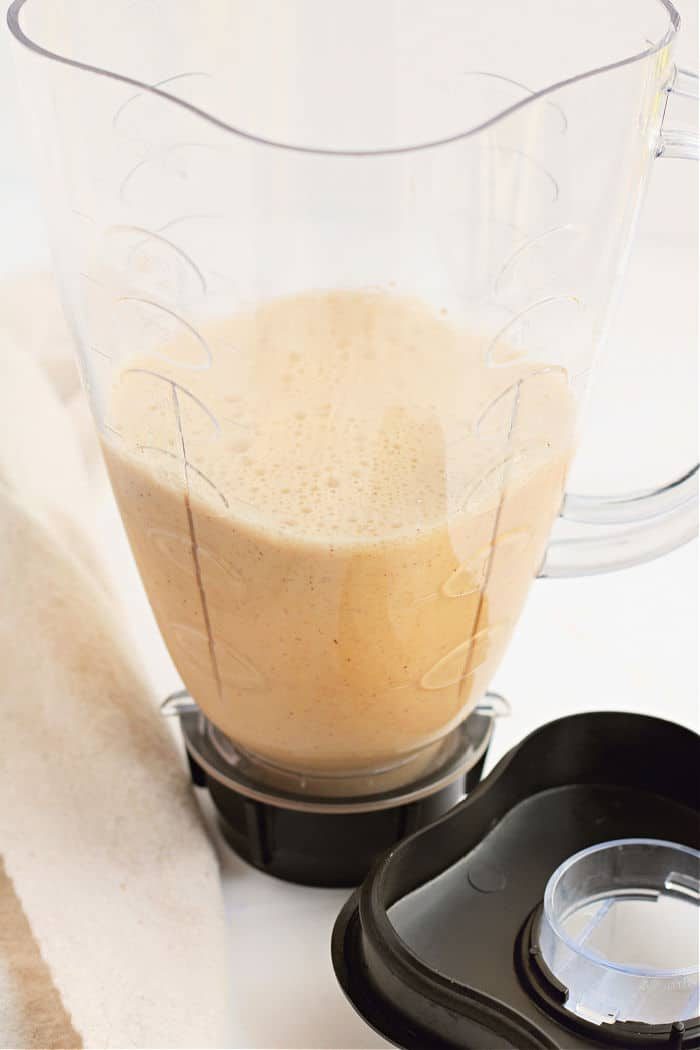 A blender pitcher filled with a creamy, light brown smoothie, reminiscent of pancakes in the oven, sits next to its black lid on a white surface.