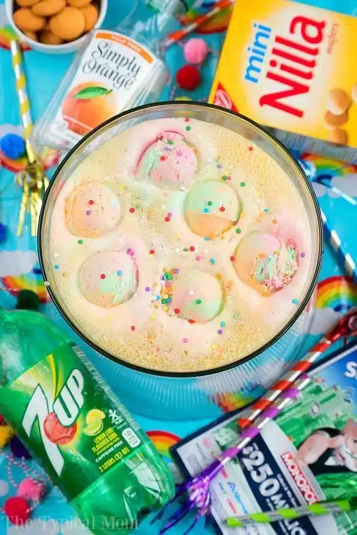 A bowl of colorful party punch with scoops of rainbow sherbet and sprinkles is surrounded by cans of Orange and 7Up, a box of Mini Nilla wafers, and packets of Kool-Aid. Straws and festive napkins add the solid touch to this delightful setup.
