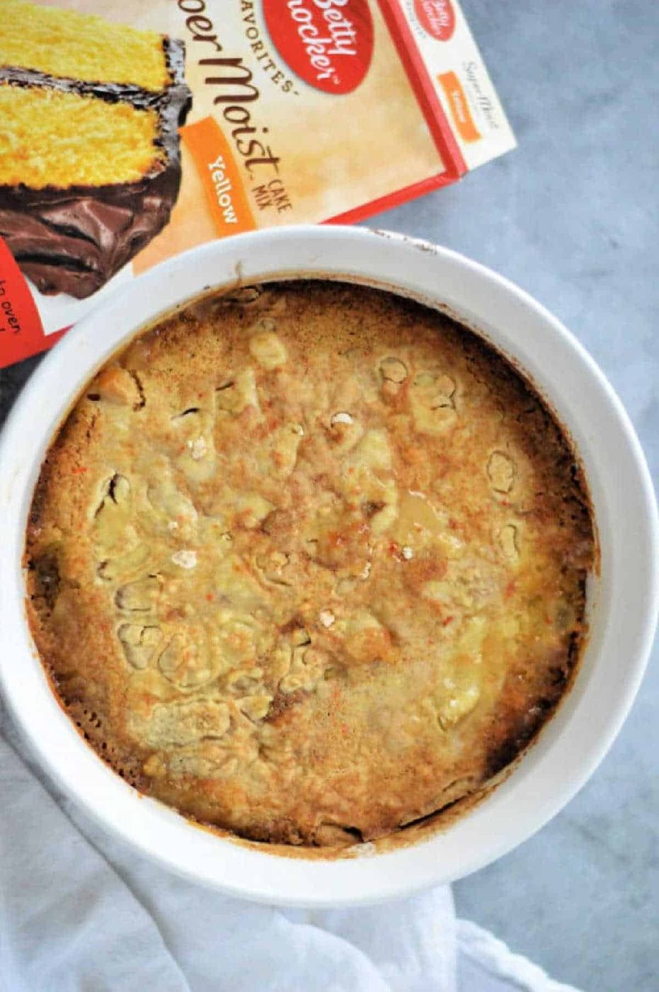 A peach cobbler with cake mix is baked in a white dish, sitting next to a Betty Crocker yellow cake mix box on a gray surface.