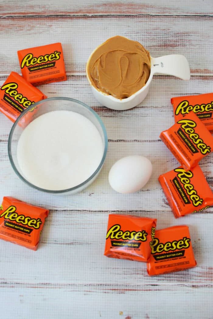 A bowl of sugar, creamy peanut butter, an egg, and several Reese's peanut butter cups are arranged on a white surface—solid for making irresistible peanut butter cookies.