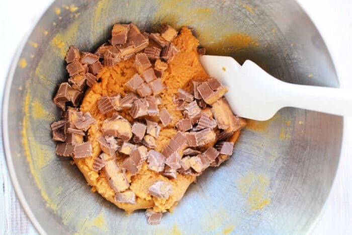 Cookie dough with chopped chocolate candy pieces and bits of Peanut Butter Cups, stirred together with a white spatula in a metal mixing bowl—solid for making the ultimate Peanut Butter Cookies.
