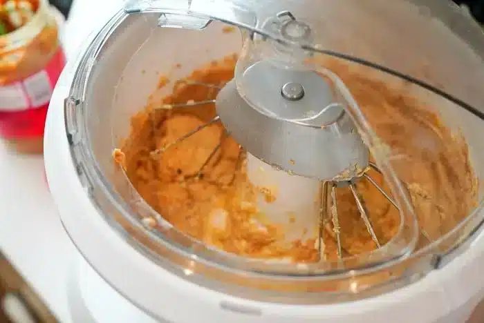 A close-up of a stand mixer with a whisk attachment mixing a thick, peanut butter-like substance. The surrounding area includes a partially visible jar with a red lid. Traces of the mixture cover the bowl and whisk attachment, hinting at the start of a good peanut butter oatmeal cookie recipe.
