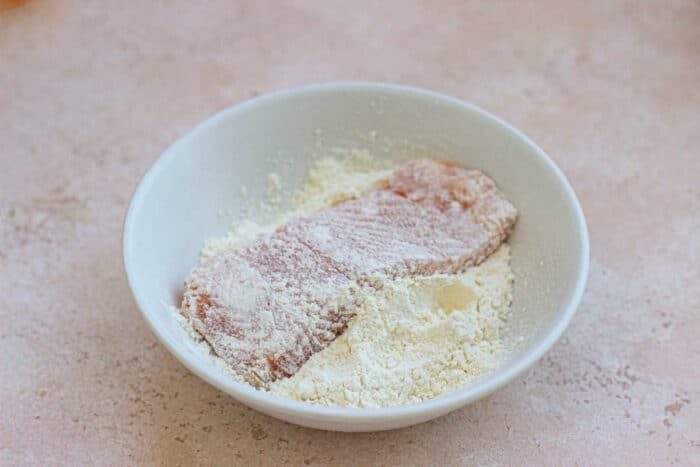 Raw trout coated in flour sits in a white bowl on a beige countertop, reminiscent of preparing pecan-crusted trout.