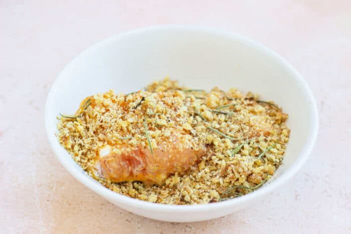 A white bowl filled with seasoned breadcrumbs and a fish fillet sits on a light surface, hinting at the nutty flavor of a pecan-crusted trout experience.