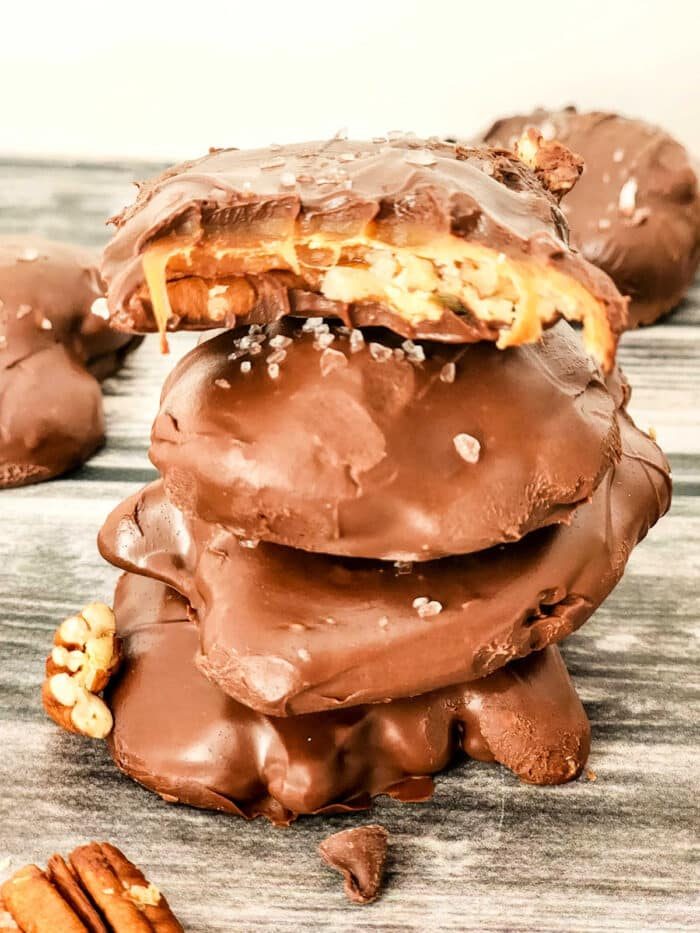 A stack of chocolate-covered caramel and pecan turtles is shown. The top cluster has a bite taken out of it, revealing luscious caramel and crunchy pecans. They are sprinkled with sea salt and sit invitingly on a wooden surface.