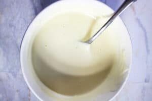 A bowl of creamy white peppermint frosting batter with a spoon resting inside on a marble surface.