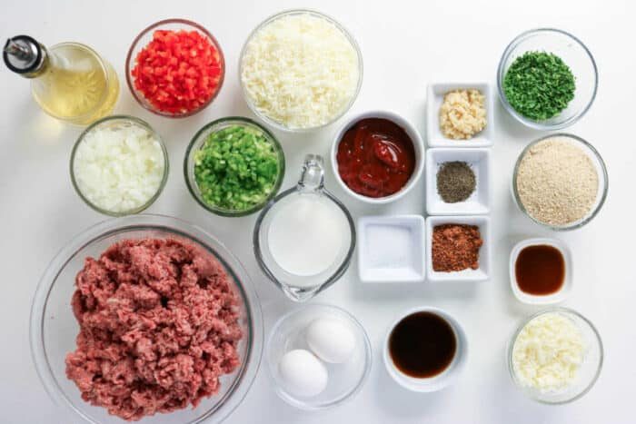 Ground meat, eggs, vegetables, seasonings, cheese, breadcrumbs, milk, and sauces arranged on a white surface—everything you need to craft a good Philly Cheesesteak Meatloaf bursting with classic flavors.