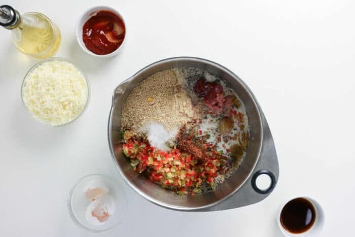 Metal bowl with eggs, breadcrumbs, diced peppers, and seasonings—solid for mixing up a good Philly Cheesesteak Meatloaf. Cheese, ketchup, and sauces are arranged in small bowls nearby to enhance the classic flavors of this meatloaf-inspired dish.