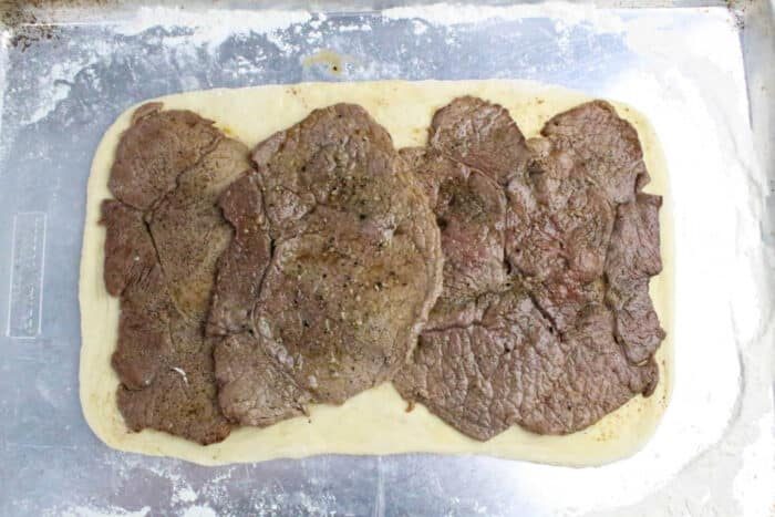 Rectangular dough topped with several overlapping slices of cooked beef, ready to be transformed into a good Philly Cheesesteak Stromboli on a floured baking sheet.