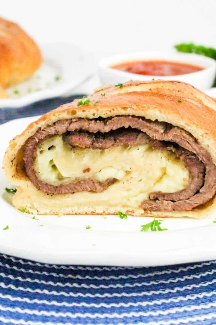 A sliced Philly Cheesesteak Stromboli with layers of beef and cheese on a white plate, sauce in the background.