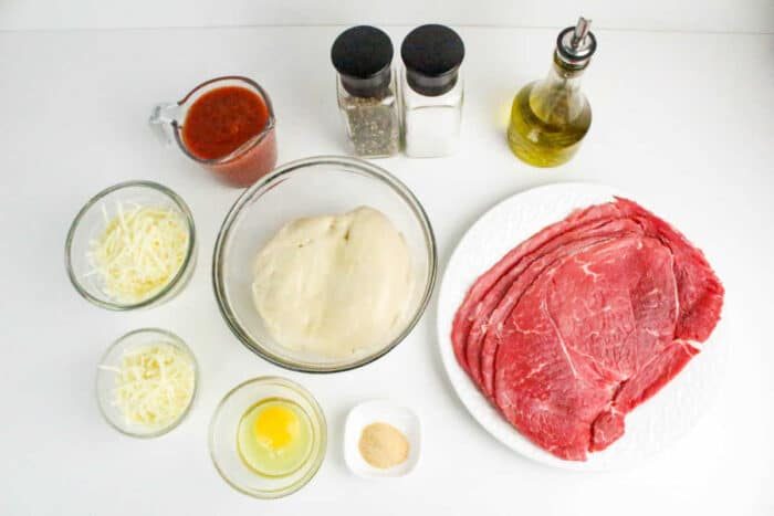 Philly Cheesesteak Stromboli ingredients: pizza dough, raw steak slices, cheese, egg, marinara sauce, oil, salt, pepper, and seasoning arranged on a white surface.