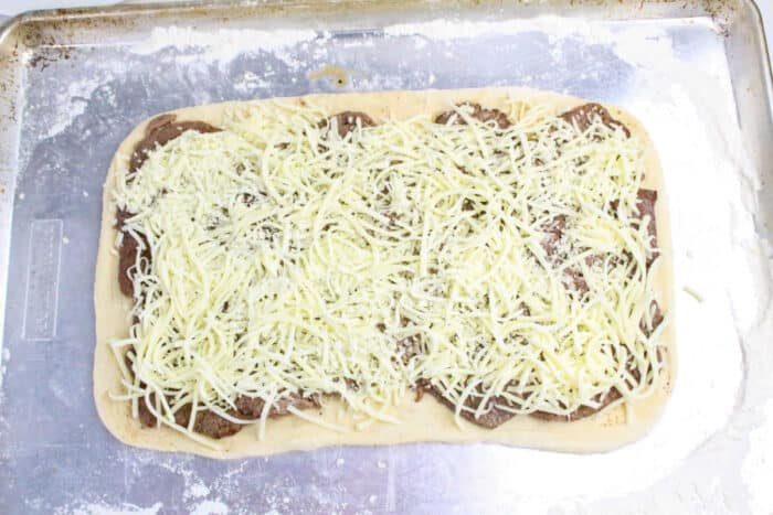 Rectangular pizza dough topped with meat slices and shredded cheese on a floured baking sheet, solid for making Philly Cheesesteak Stromboli.