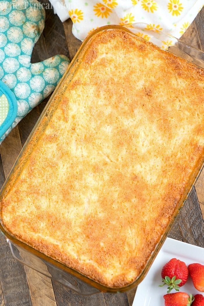 A golden-brown baked pineapple angel food cake recipe, rests in a glass dish. Nearby, a blue oven mitt and a plate with strawberries are artfully arranged on a wooden table.