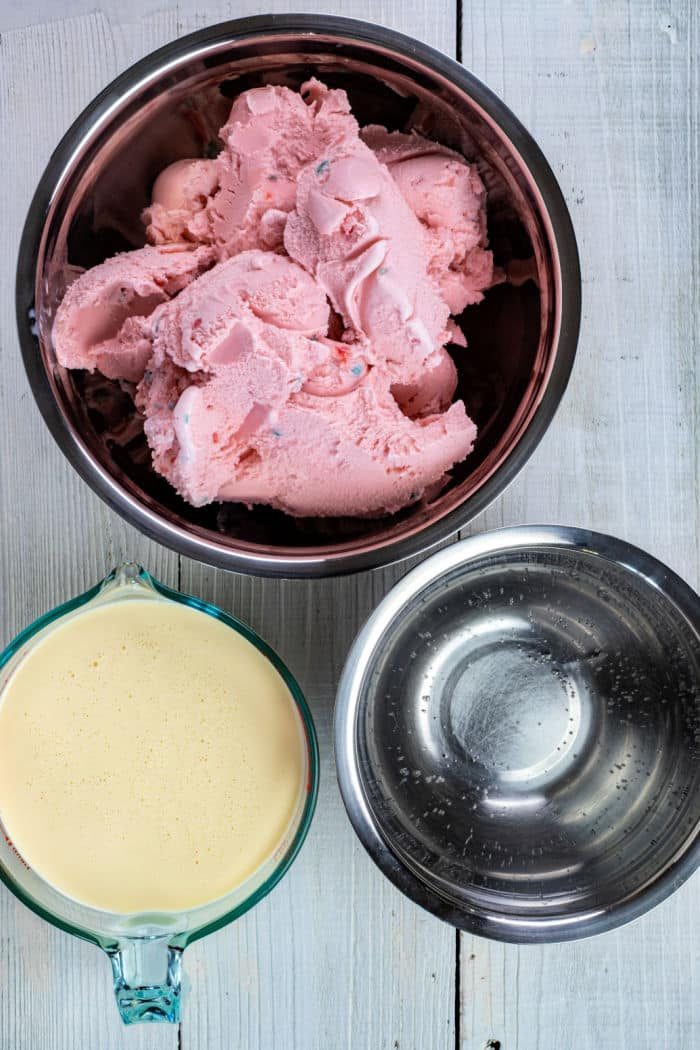 Three bowls on a white surface: one with pink punch recipe-inspired ice cream, one with custard, and one empty metal bowl.