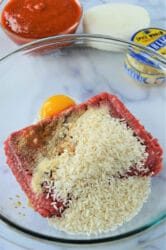 A glass bowl brimming with ground beef, an egg, uncooked rice, and spices sits on the countertop, ready to transform into instant pot porcupine meatballs. In the background, a bowl of red sauce accompanies an onion and a jar of minced garlic.
