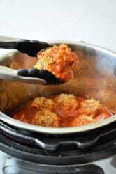 Tongs holding a portion of porcupine meatballs in front of an Instant Pot filled with stuffed peppers in tomato sauce. The dish consists of ground meat and rice in red sauce.