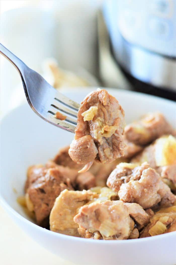 A fork holds a piece of tender pork adobo above a bowl filled with chunks of meat and onions, freshly made in an Instant Pot.