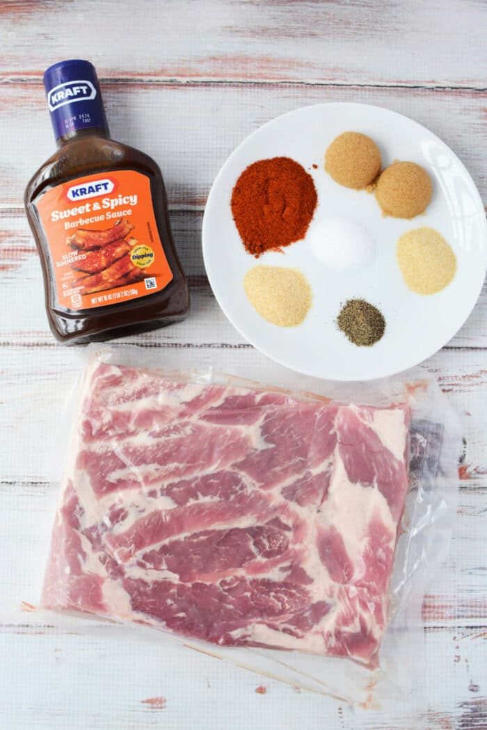 A slab of raw pork ribs, barbecue sauce, and a plate of six different spices await your next recipe on a white wooden surface—solid for crafting smoky burnt ends or experimenting with pork belly delights.