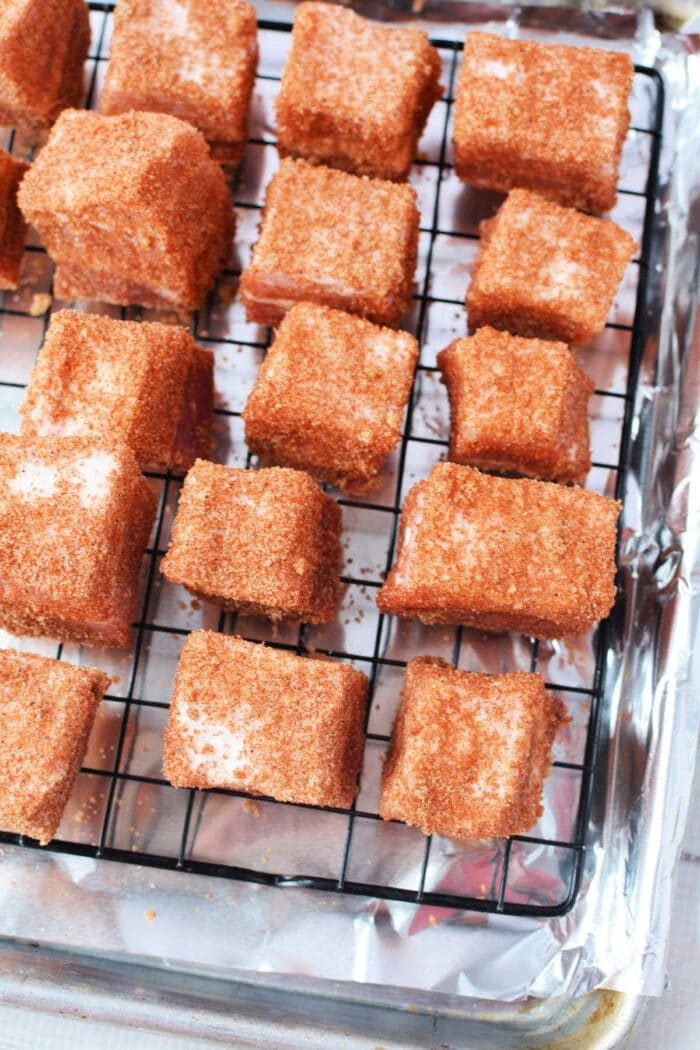 Squares of pork belly coated in a reddish-brown seasoning rest on a wire rack over a foil-lined tray, solid for a savory burnt ends recipe.