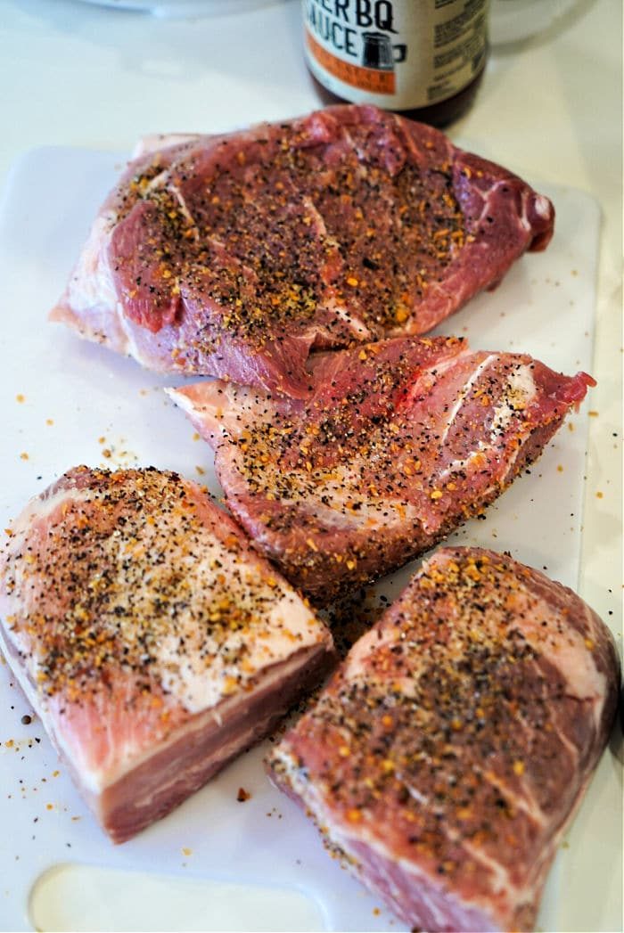 Close-up of seasoned raw boston butt on a white cutting board beside a jar of BBQ sauce, reminiscent of the savory aromas when preparing pork butt in an Instant Pot.