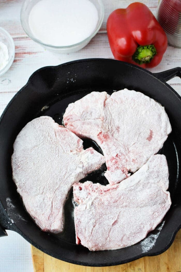 Three flour-coated raw pork chops in a black skillet, with a red bell pepper and a bowl nearby—solid ingredients for trying new pork chop recipes.