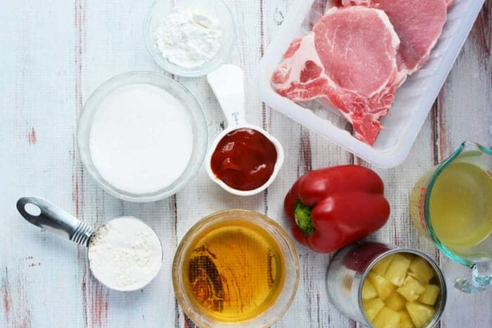 Raw pork chops, red bell pepper, flour, sugar, ketchup, pineapple, broth, and cornstarch sit on a white surface—ideal ingredients for good Pork Chop Recipes.