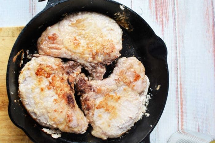 Four pork chops cooking in a black cast iron skillet on a light wooden surface, solid for a savory pork and pineapple meal.