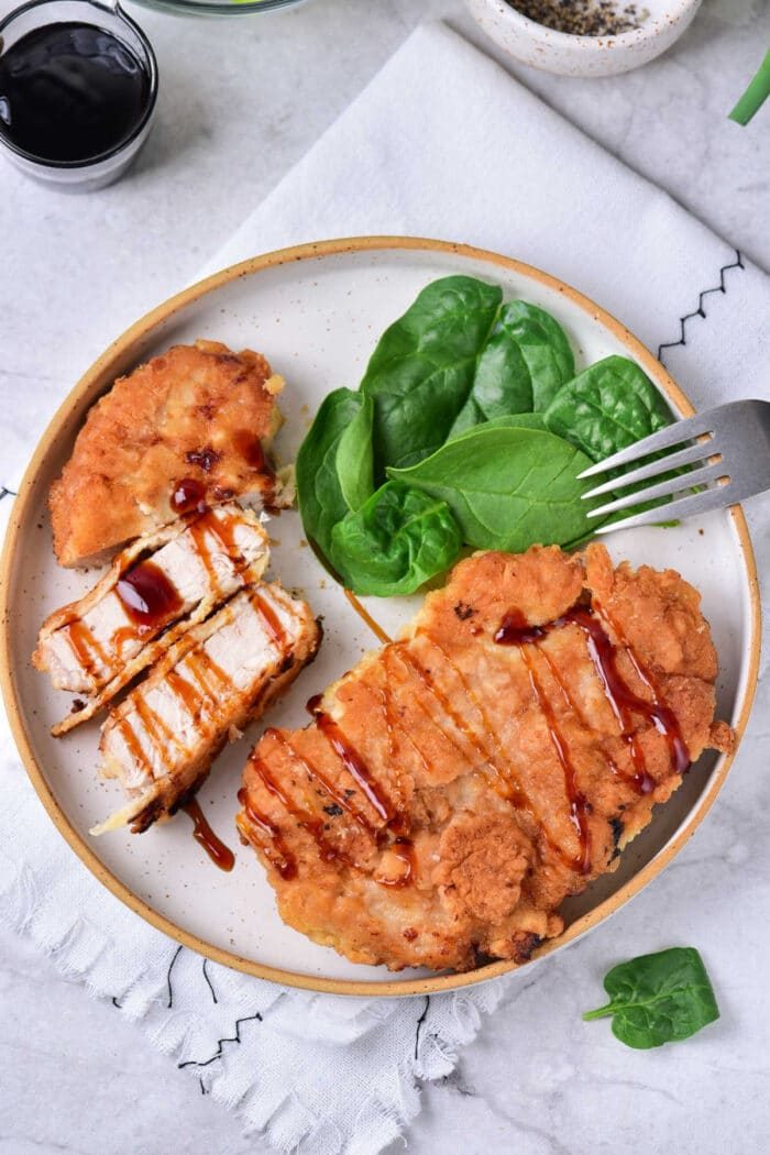Fried pork cube steak drizzled with barbecue sauce rest alongside fresh spinach and a fork on a plate.