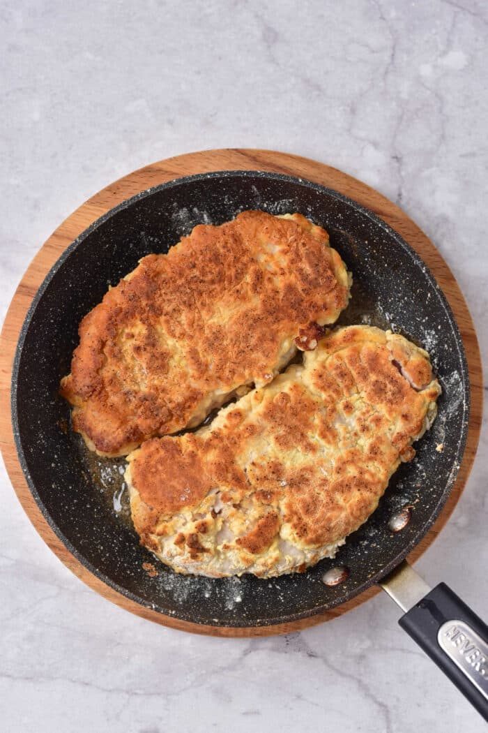 Two breaded pork cutlets and a succulent pork cube steak sizzle in a black pan set on a wooden board.