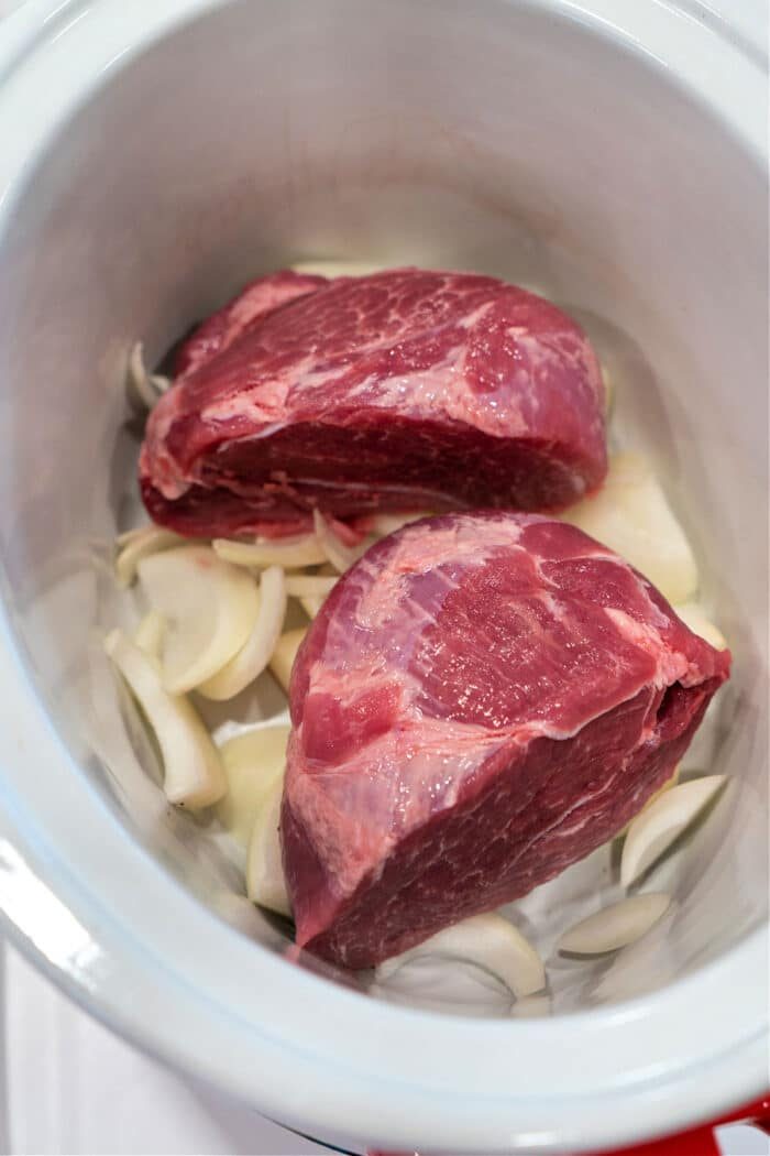 Two raw pork picnic roast rest in a slow cooker, surrounded by sliced onions.