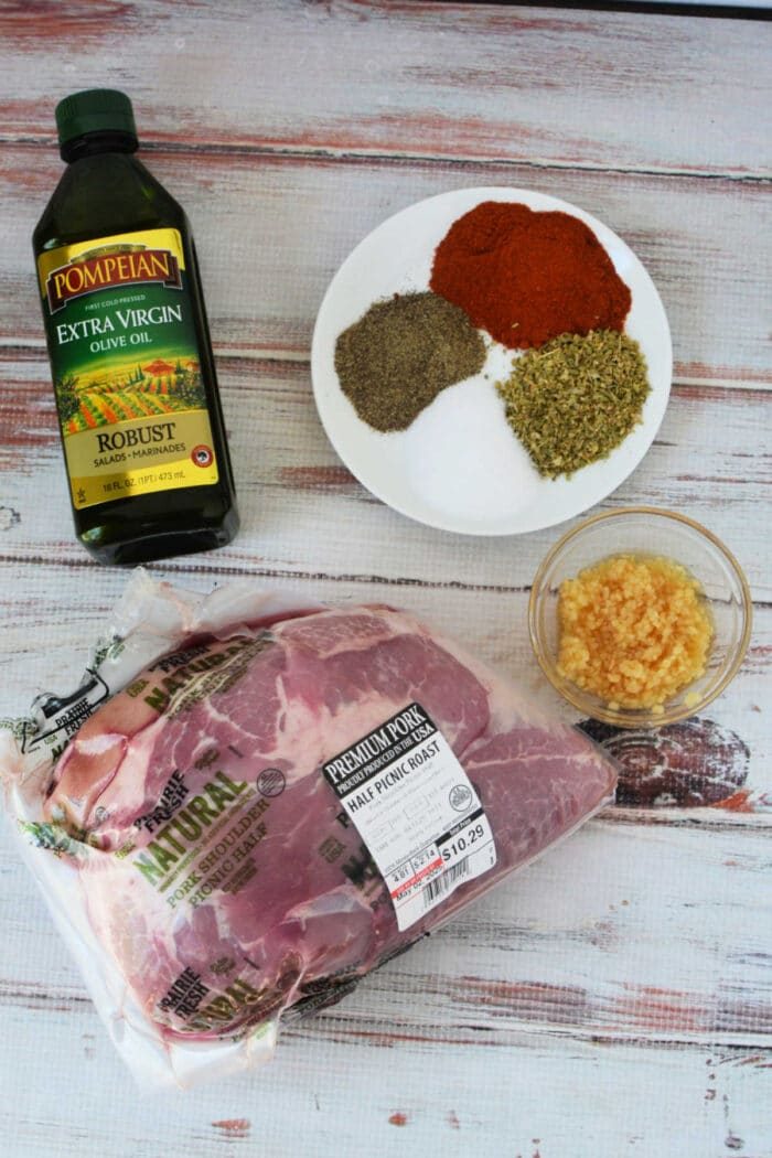 Pork picnic Roast Recipe, with a bottle of olive oil, minced garlic, and a plate of herbs and spices on a white wood surface—solid ingredients before roasting in the oven.